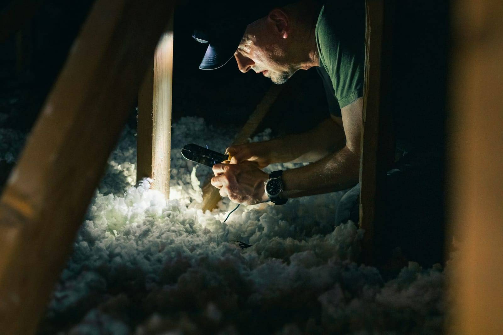 A man in a cap working with pliers by the light of a headlamp in a dark attic, between wood framing and loose-fill insulation