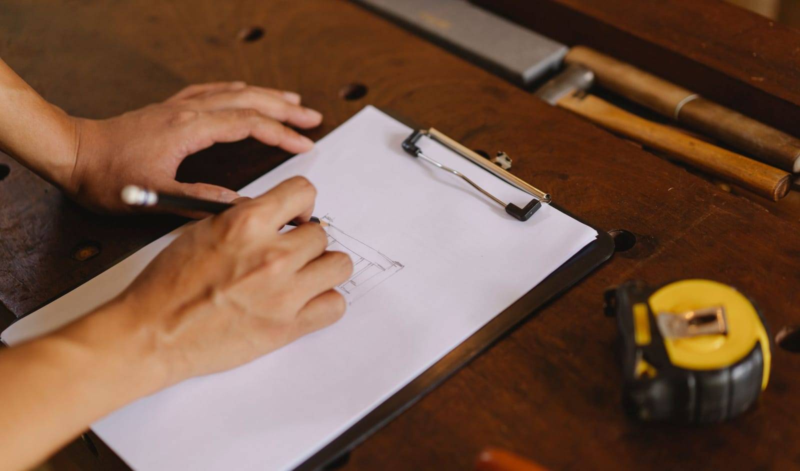 Hands writing on a clipboard on a wooden workbench, with a tape measure and hand tools nearby