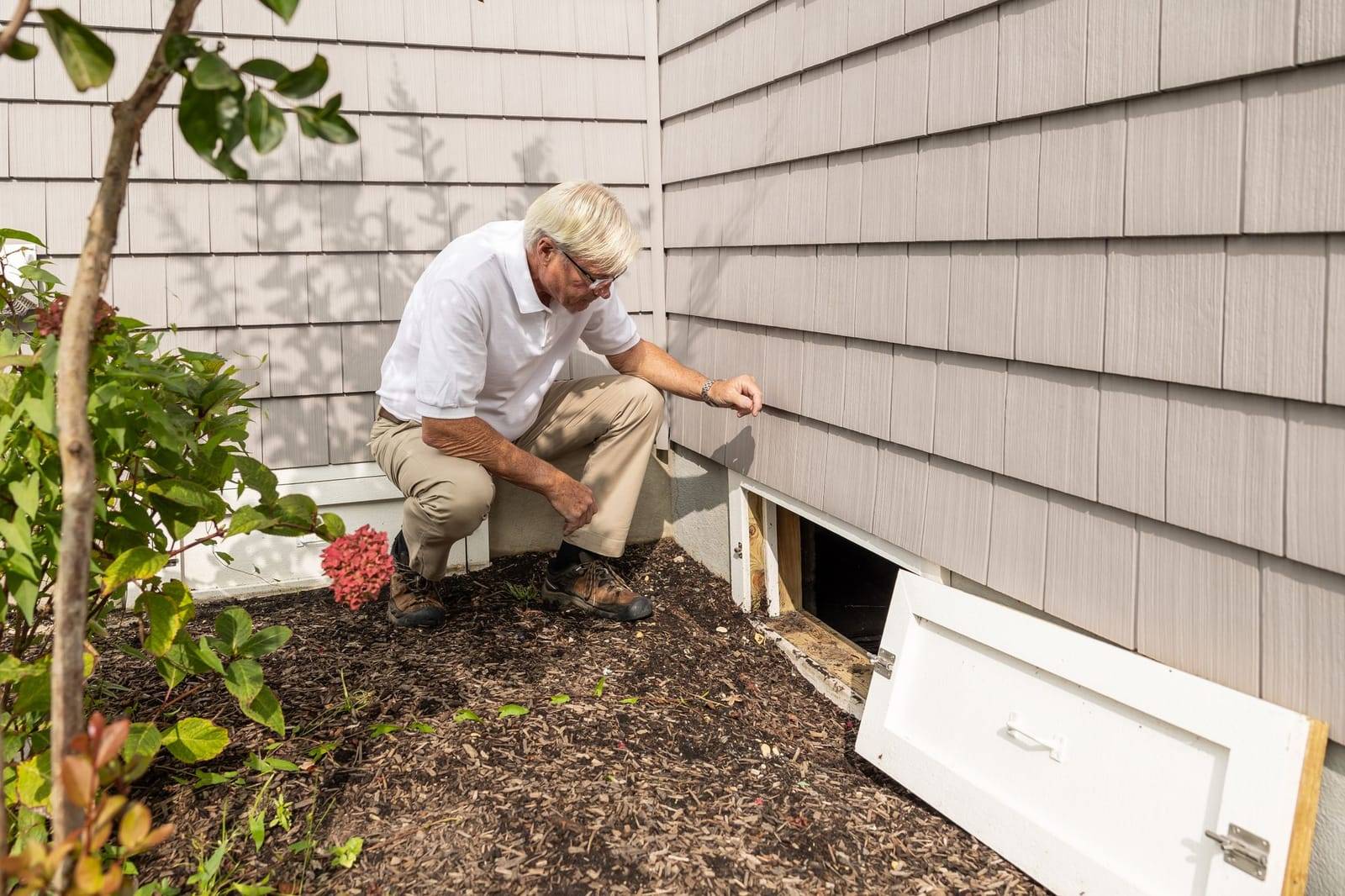 A man crouching beside the open crawl space access door at the base of a house wall, looking inside
