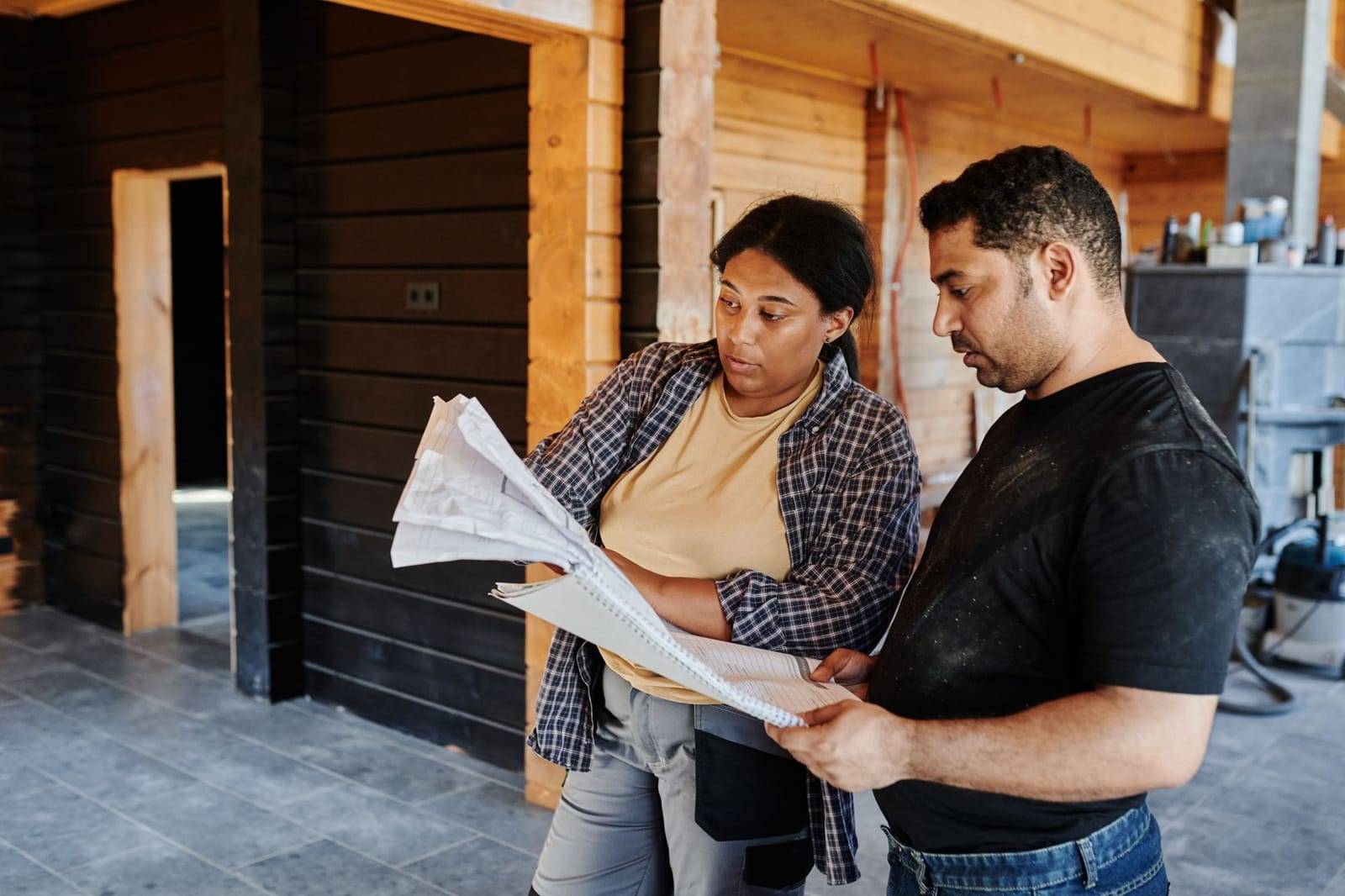 A woman and a man in work clothes reading printed plans together inside a house under construction